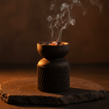 Matte black bakhoor burner with glowing coal and incense smoke on dark stone
